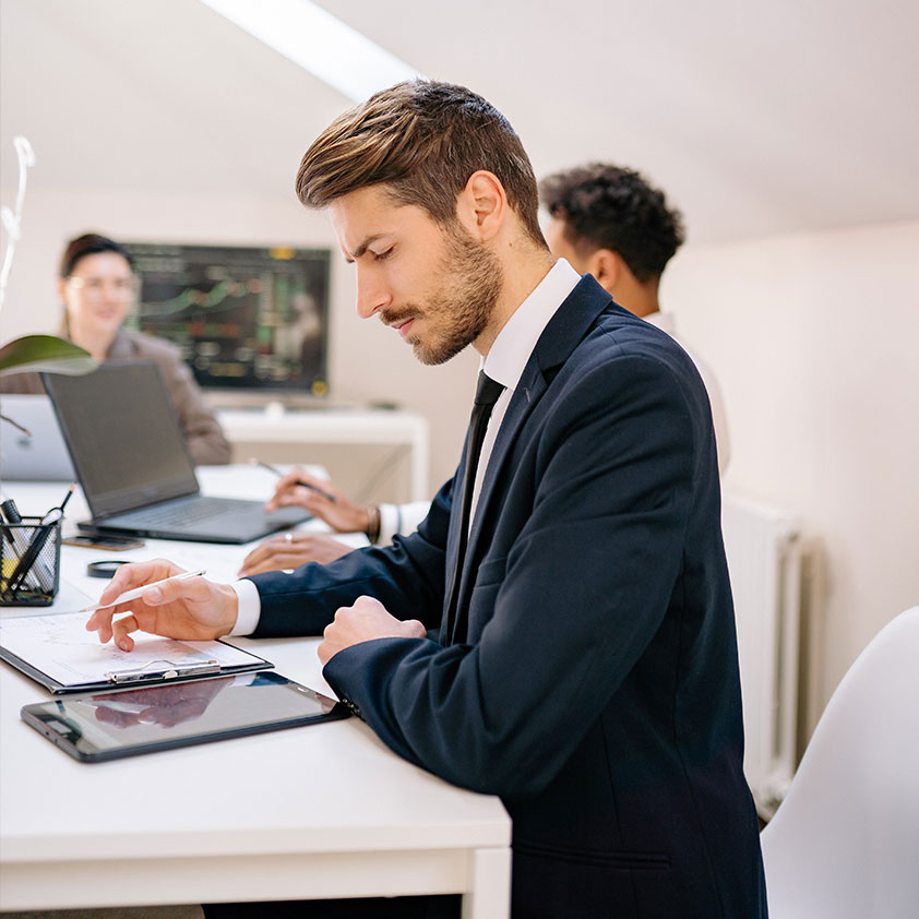 Business Consultant taking notes Man in suit sitting in front of a notebook working, women in the background. Office Setting.